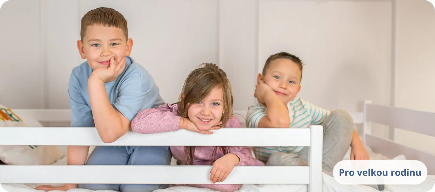 Tři usmívající se děti opírající se o bílou zábranu postele.